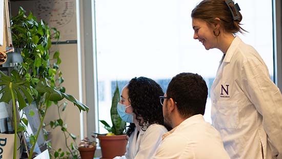 A Northwestern researcher in a white lab coat stands behind two researchers seated at a computer to support them.